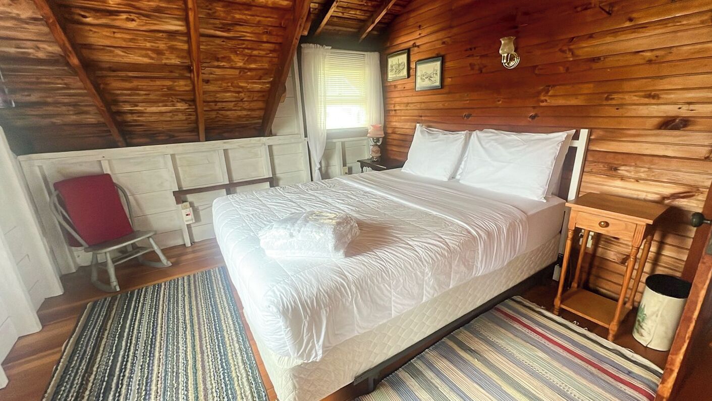 A wooden cabin bedroom with a bed, a chair, a nightstand, and a window with curtains.