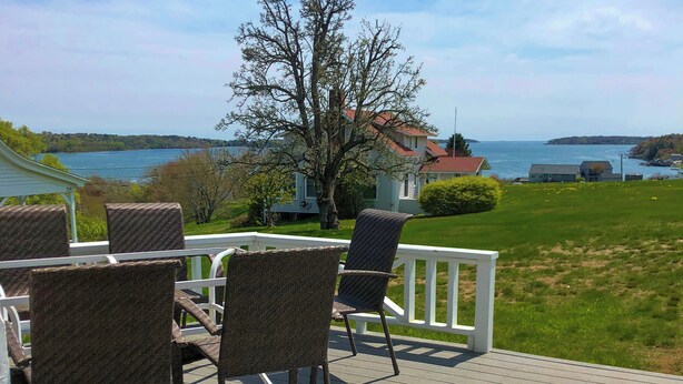 A deck with wicker chairs and a table, overlooking a lake with houses in the background. - Thorburn - Experience a classic ME vacation at this beautiful summer cottage (Harpswell)
