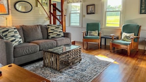 A cozy living room with a wooden ceiling, a gray sofa with patterned cushions, a wooden coffee table, and a green armchair. - Thorburn - Experience a classic ME vacation at this beautiful summer cottage (Harpswell)