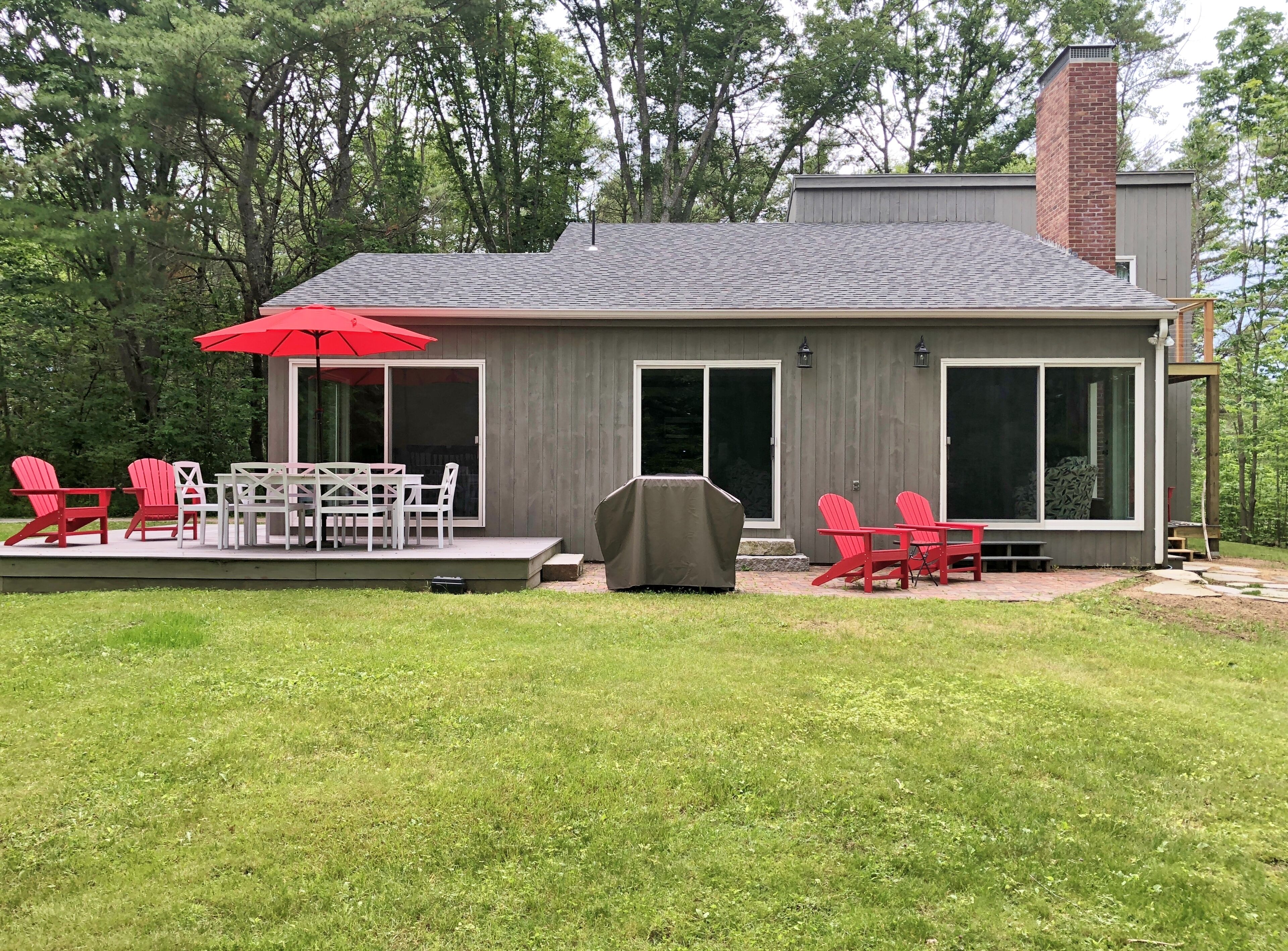 Une maison moderne dotée d’un patio comprenant une table et des chaises, un barbecue couvert et un parasol rouge.