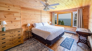 Un dormitorio de cabaña de madera con cama, cómoda, mesita de noche y vista al océano.