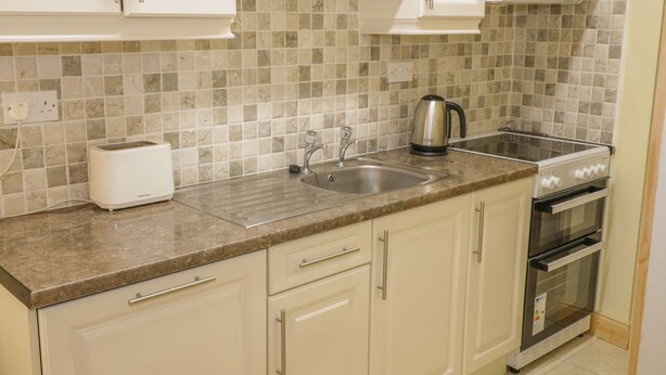A kitchen with white cabinets, a granite countertop, a stainless steel sink, a kettle, and a stove. - Glebe House (Ballinrobe)