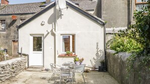 A small, single-story house with a white exterior, a front door, and a window. There is a small patio area with a table and chairs, and a potted plant on the windowsill. - Glebe House (Ballinrobe)