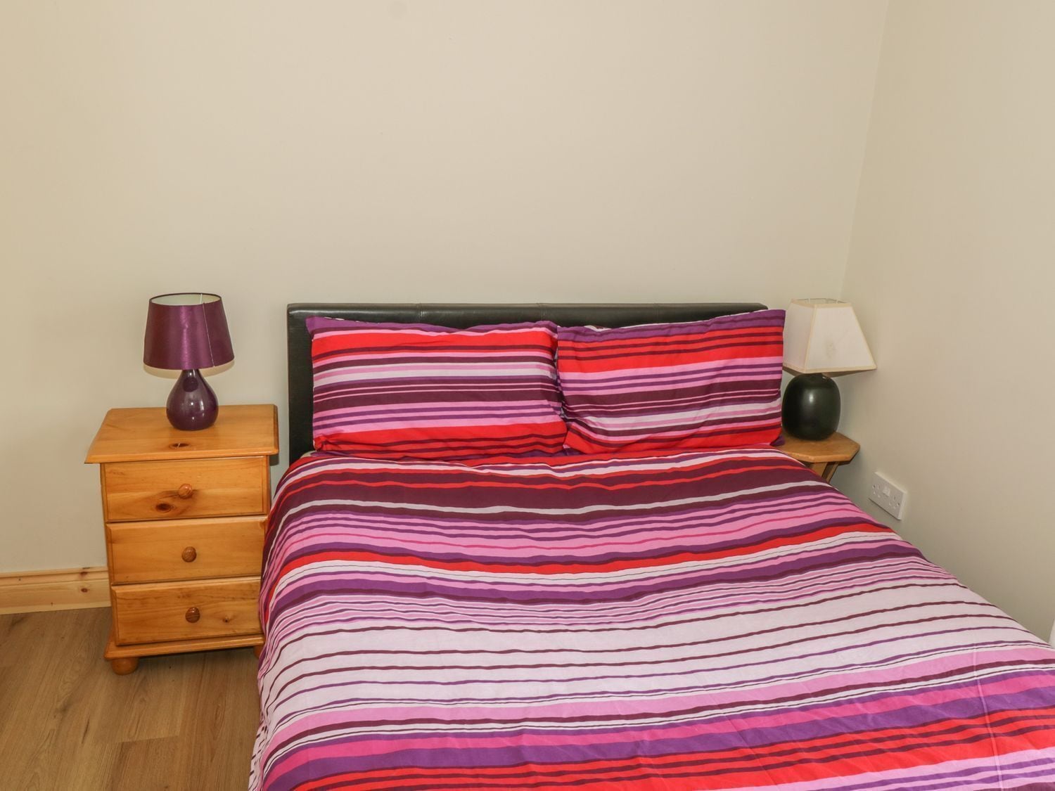 A bed with a striped bedspread, a wooden nightstand with a lamp, and a drawer unit.