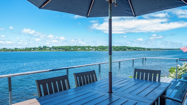 Une terrasse en bois avec une table et des chaises, donnant sur une étendue d’eau dont on aperçoit la rive au loin.