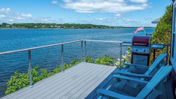 A deck with a grill and a flag, overlooking a lake with a distant shoreline. - Sunset Cottage - Capture Breathtaking Sunsets from this Beautiful Home (Bailey Island)