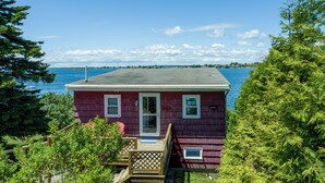 A red wooden house with a deck, a door, and a window, overlooking a body of water. - Sunset Cottage - Capture Breathtaking Sunsets from this Beautiful Home (Bailey Island)