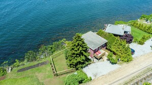 Une maison avec une terrasse et un grand arbre à l’avant, située près d’une étendue d’eau.