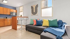 A modern living room with a gray sofa, colorful cushions, a kitchen area with wooden cabinets and a microwave, and a large window with a view. - Puerto Rico Ocean Park Beach Vacation Rental Otium Rentals (San Juan)