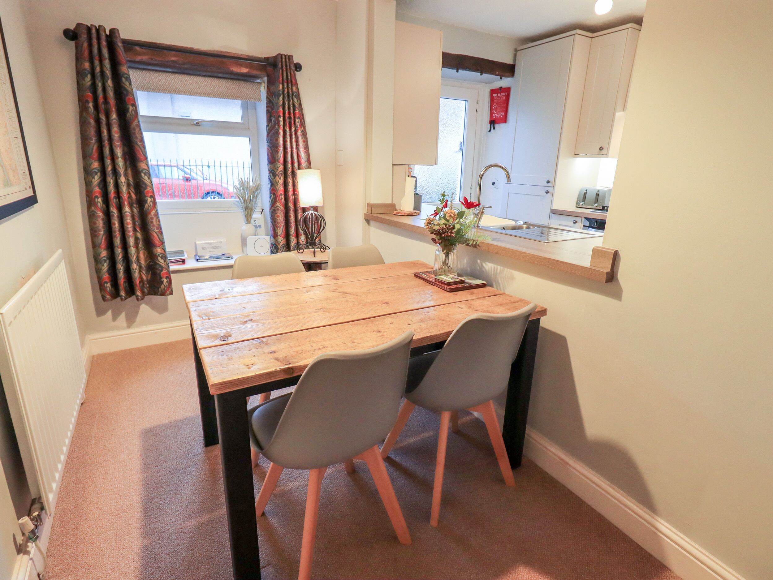 A dining area with a wooden table, two grey chairs, a vase with flowers, and a view of the outside through a window with patterned curtains.