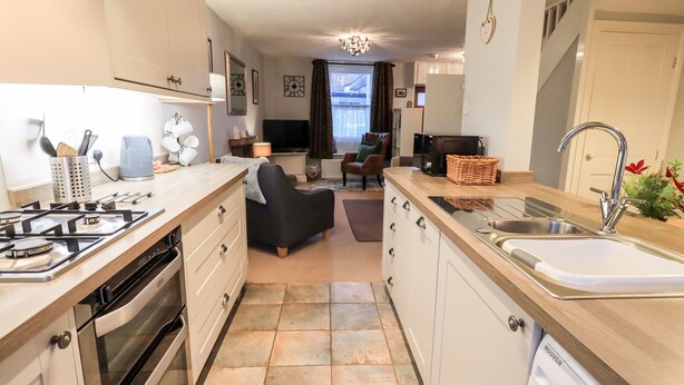 A modern kitchen with a gas stove, oven, and dishwasher. There is a dining area with a sofa and a TV in the background. - The Old Betting Shop (Keswick)