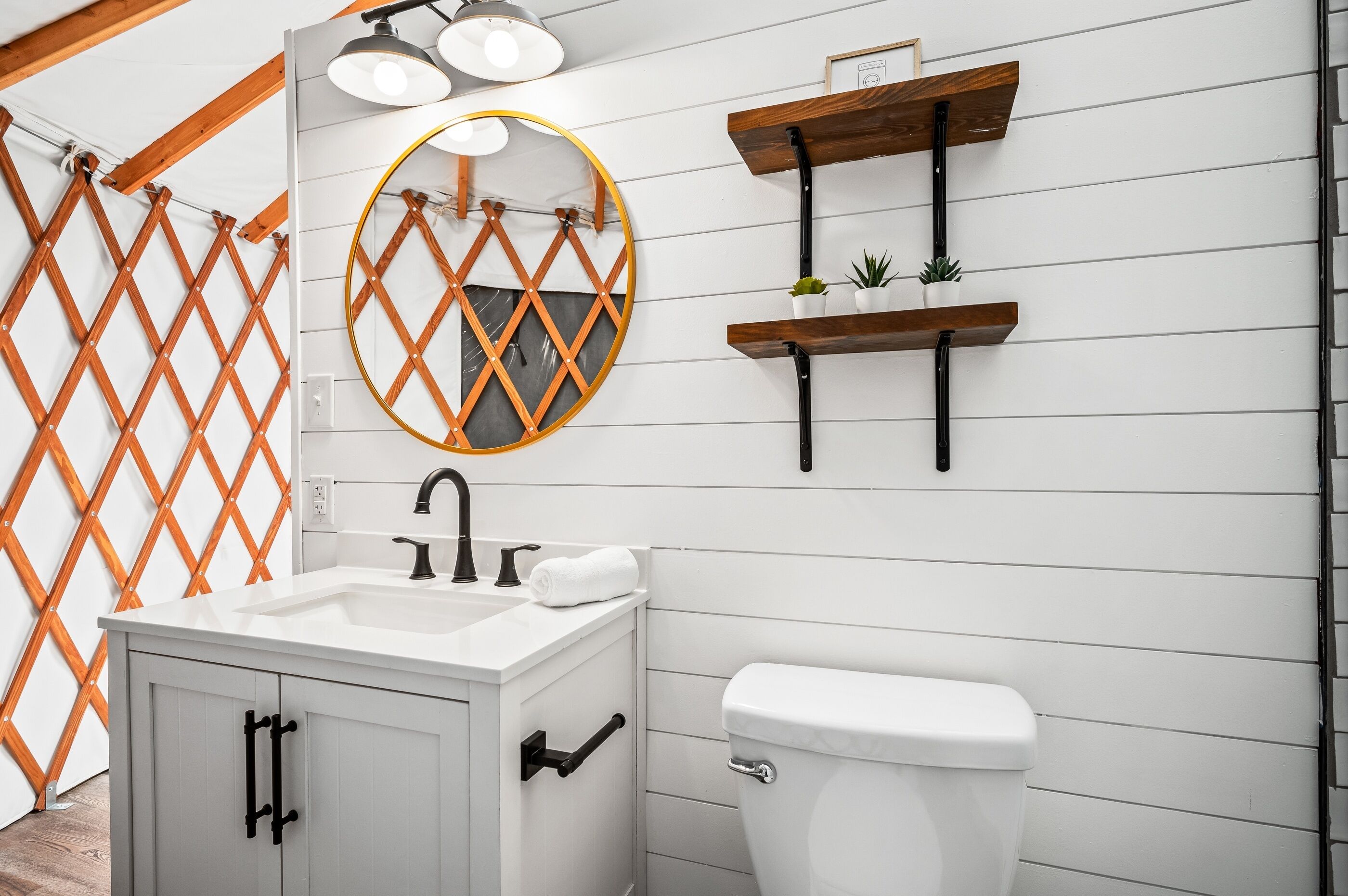 A bathroom with a white sink, a toilet, a mirror, and wooden shelves with plants.