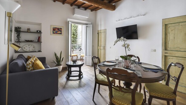 A living room with a sofa, a dining table with chairs, a TV mounted on the wall, and a wooden ceiling.