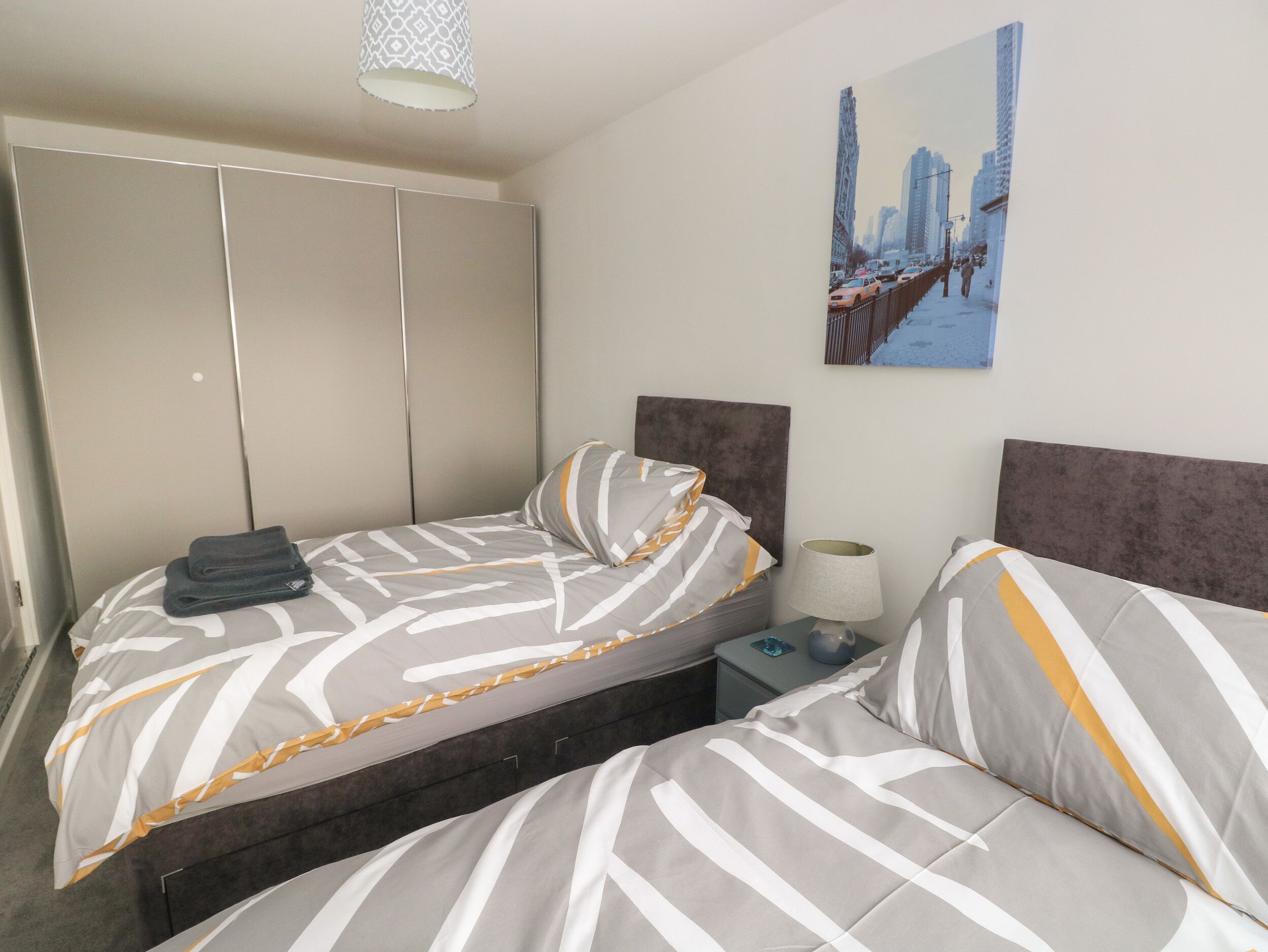 A hotel room with two beds, a wardrobe, a nightstand, and a cityscape painting.