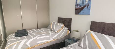 A hotel room with two beds, a wardrobe, a nightstand, and a cityscape painting.