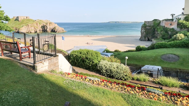 Vista costeira com praia, um forte histórico e um jardim com flores coloridas.