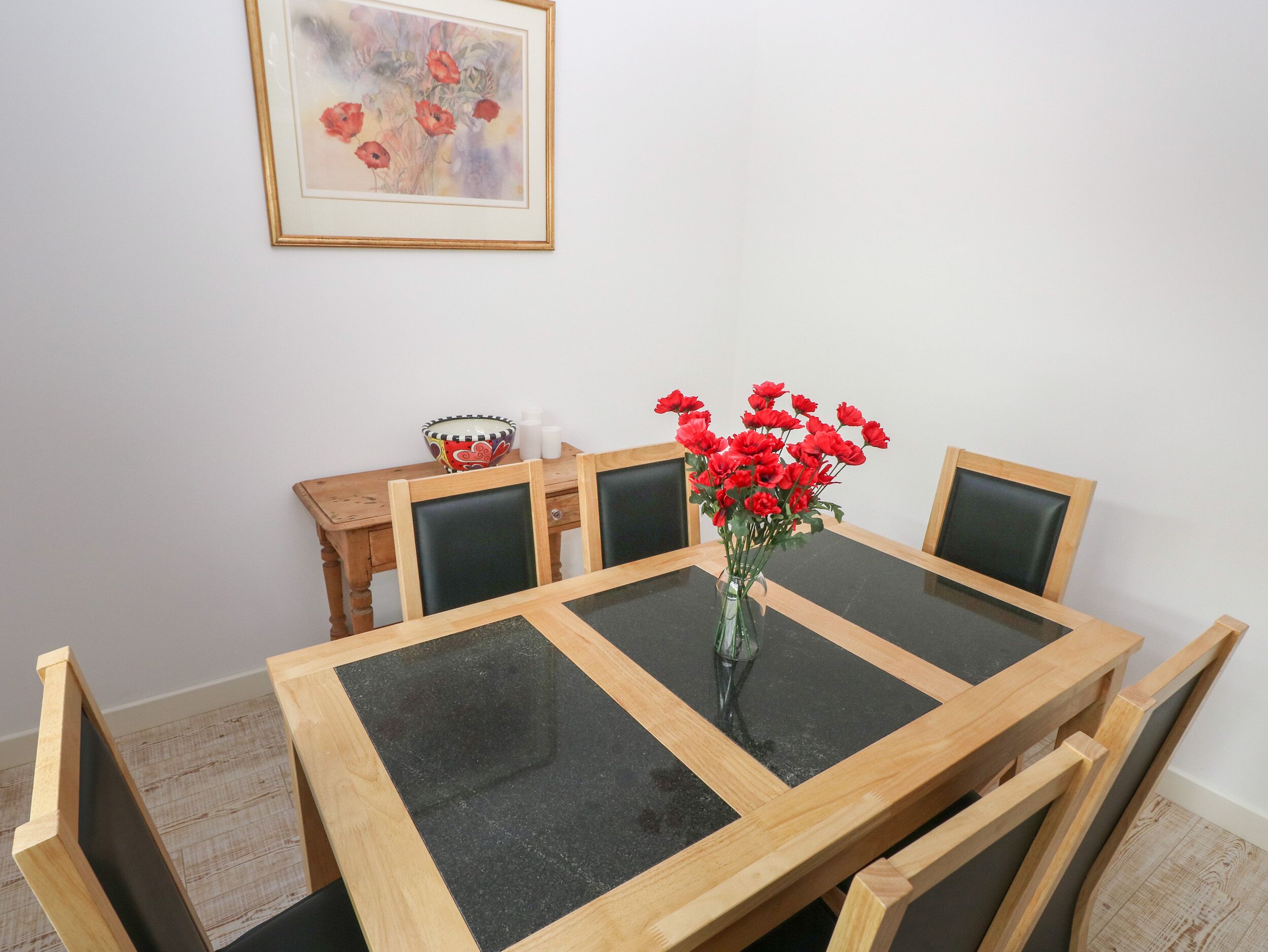 Ett matbord med en glasskiva och träram, omgivet av fyra stolar. En vas med röda blommor står på bordet.