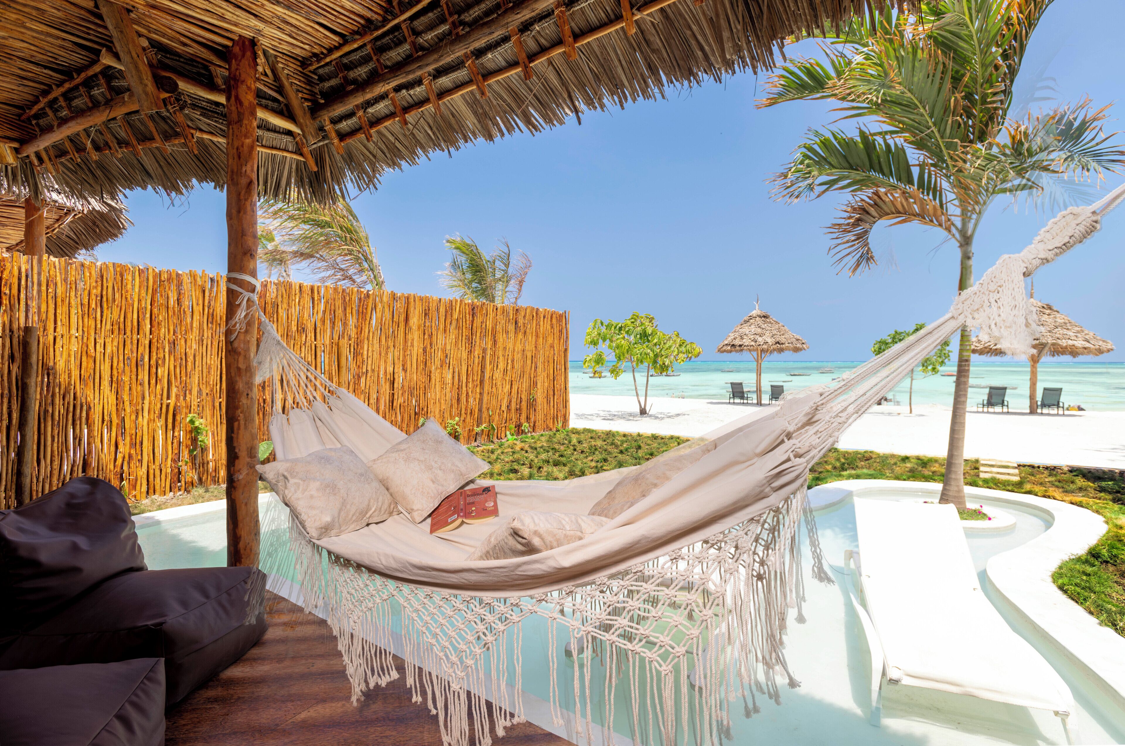 A hammock with macrame fringe, a wooden deck, a pool, and a beach view.