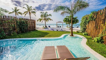 A swimming pool with lounge chairs, a bamboo fence, and a view of the beach with palm trees.