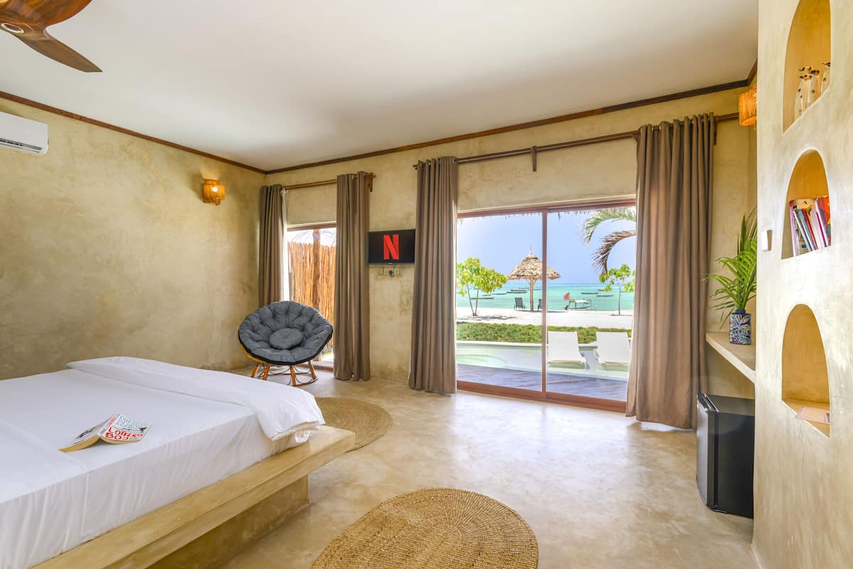 A bedroom with a bed, a chair, a TV, and a view of a pool and beach.