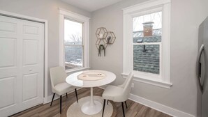 Una habitación con una mesa redonda blanca, dos sillas, un adorno en la pared de forma hexagonal y ventanas amplias con vistas a una chimenea de ladrillo.
