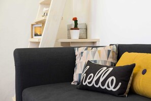 A dark grey sofa with three cushions, one with "hello" text, another with a geometric pattern, and a yellow one. A small wooden shelf with a potted plant and a framed picture is behind the sofa. - Design Apt Florence (Florence)