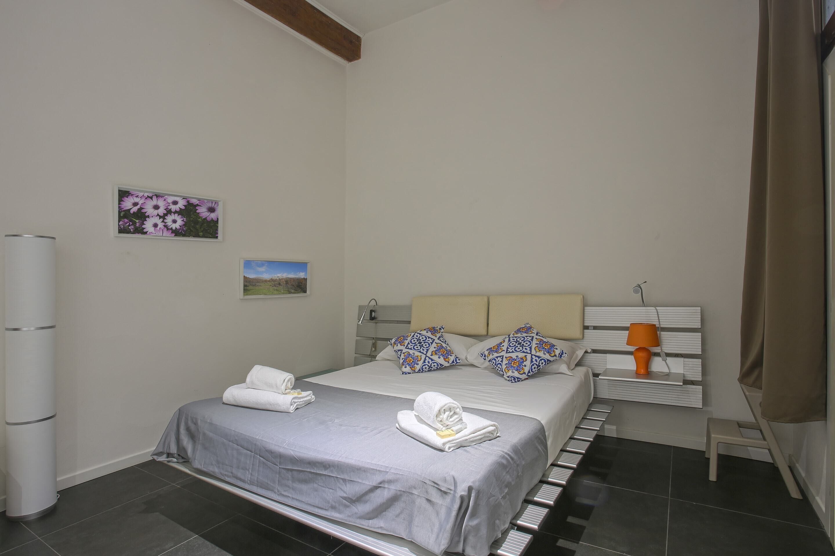 Cama bem arrumada com colcha cinza e almofadas brancas, mesa de cabeceira com abajur e prateleira na parede com porta-retrato.