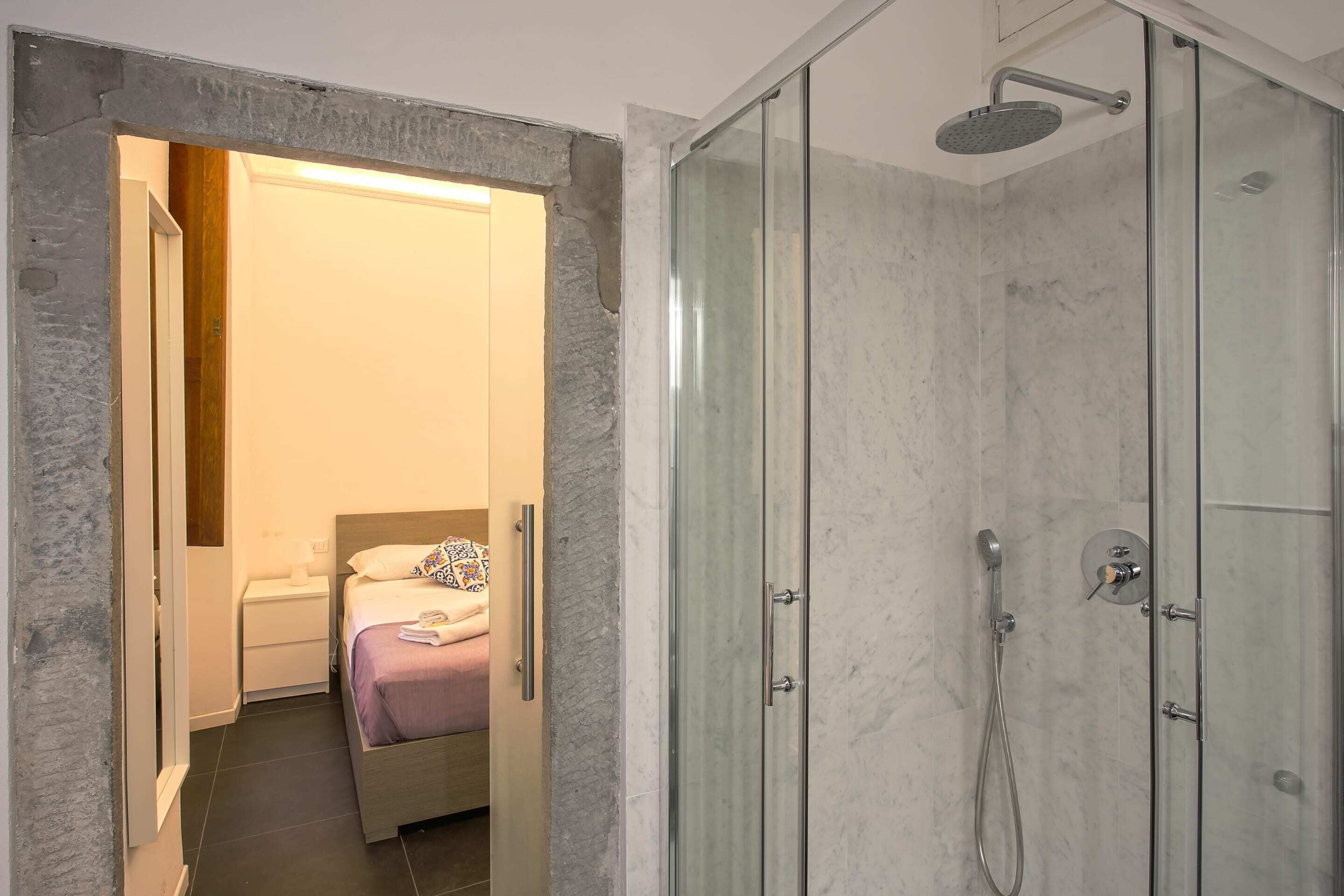 A modern bathroom with a glass-enclosed shower, a bed with a purple bedspread, and a white bedside table.