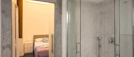 A modern bathroom with a glass-enclosed shower, a bed with a purple bedspread, and a white bedside table.