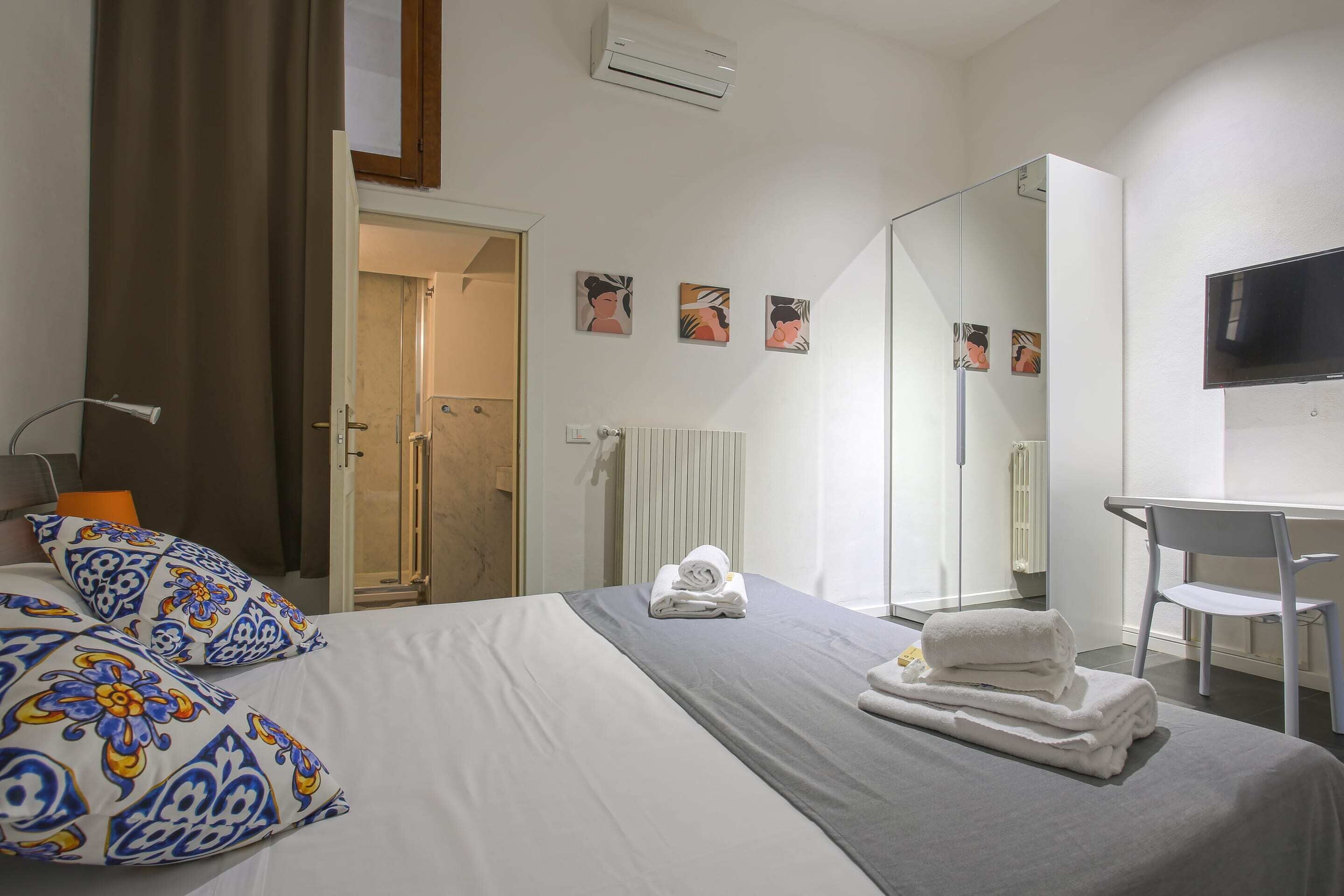 A hotel room with a bed, a desk, a chair, a TV, and artwork on the wall.