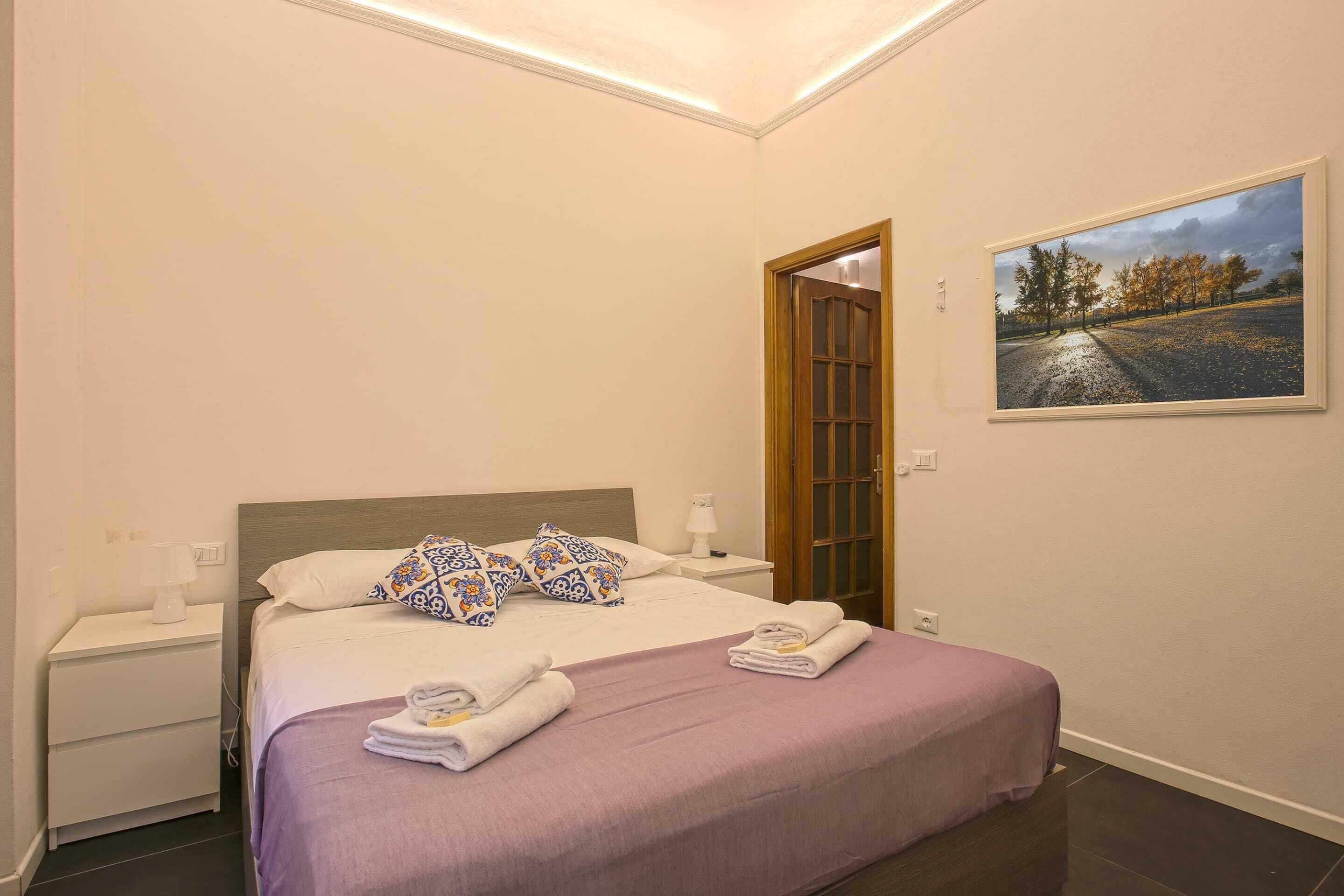A neatly made bed with a purple bedspread, white pillows, and a small bedside table with a lamp.