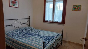A single bed with a patterned bedspread, a window with curtains, and two wall-mounted paintings. - PEPE house, cottage with garden 5 min from Campus beach, Villasimius  (Sardegna)