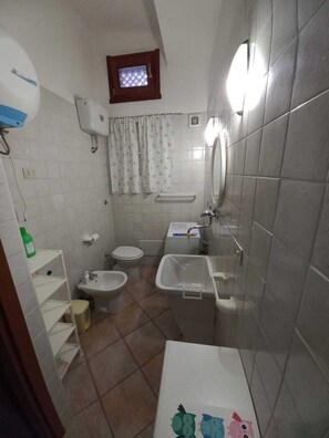 A small bathroom with a toilet, bidet, and sink. There is a window with a curtain, and a small mirror above the sink. - PEPE house, cottage with garden 5 min from Campus beach, Villasimius  (Sardegna)