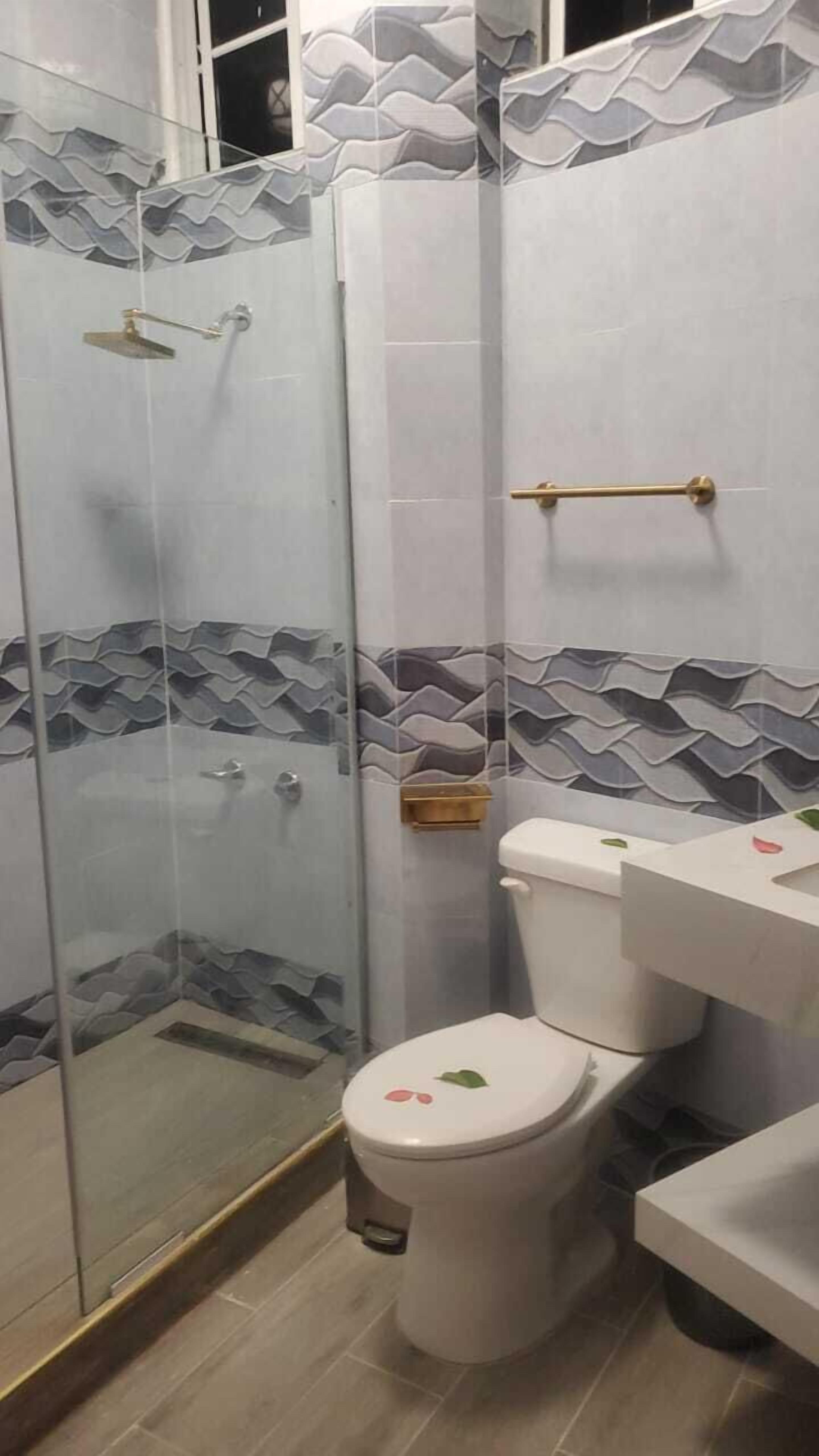 Une salle de bain dotée d’une douche à parois de verre, de toilettes dont le couvercle est orné d’un motif de feuilles et d’un lavabo avec un robinet doré.