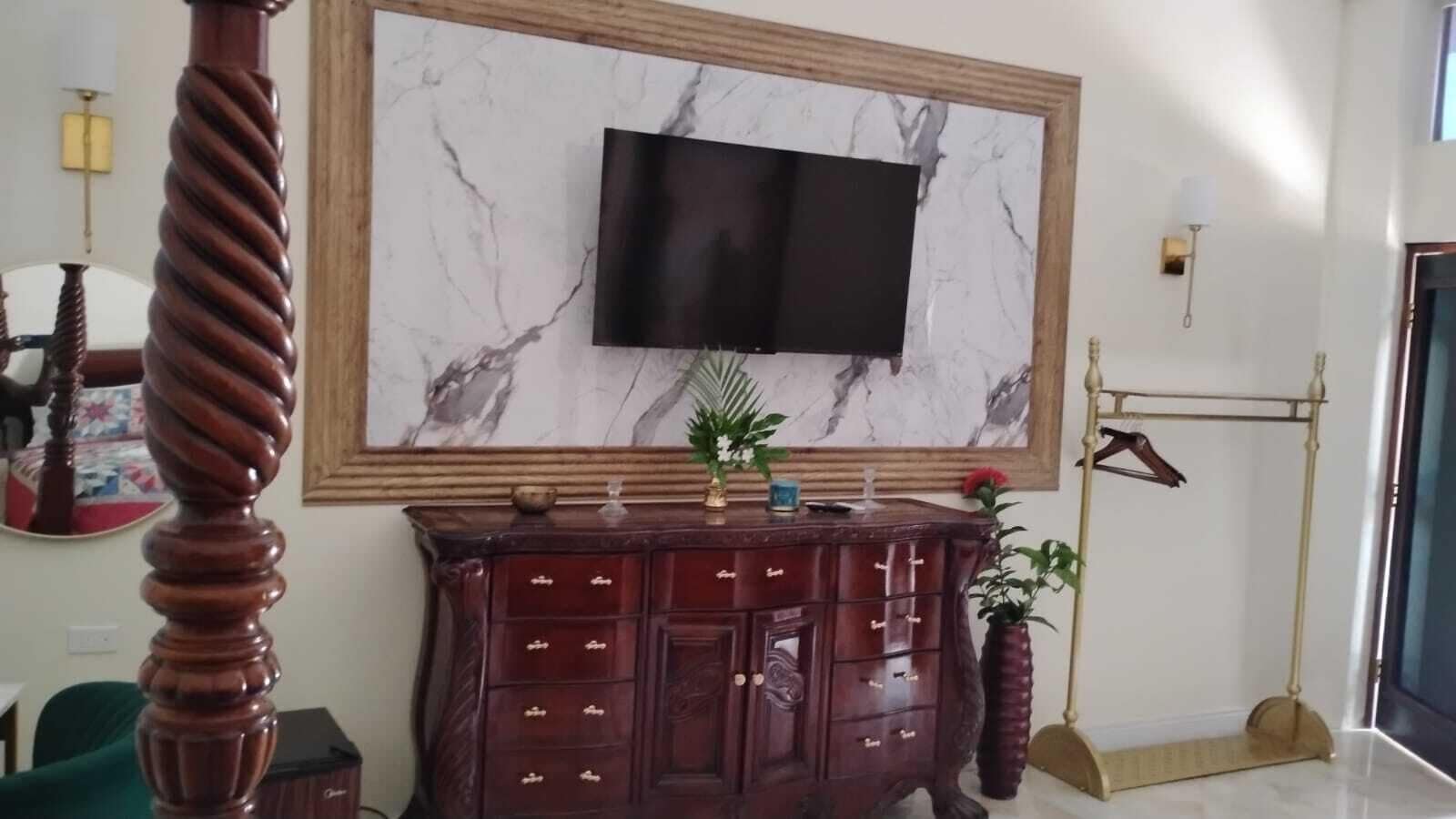 A room with a large marble wall-mounted TV, a wooden cabinet with drawers, a floor lamp, and a hanging rack.