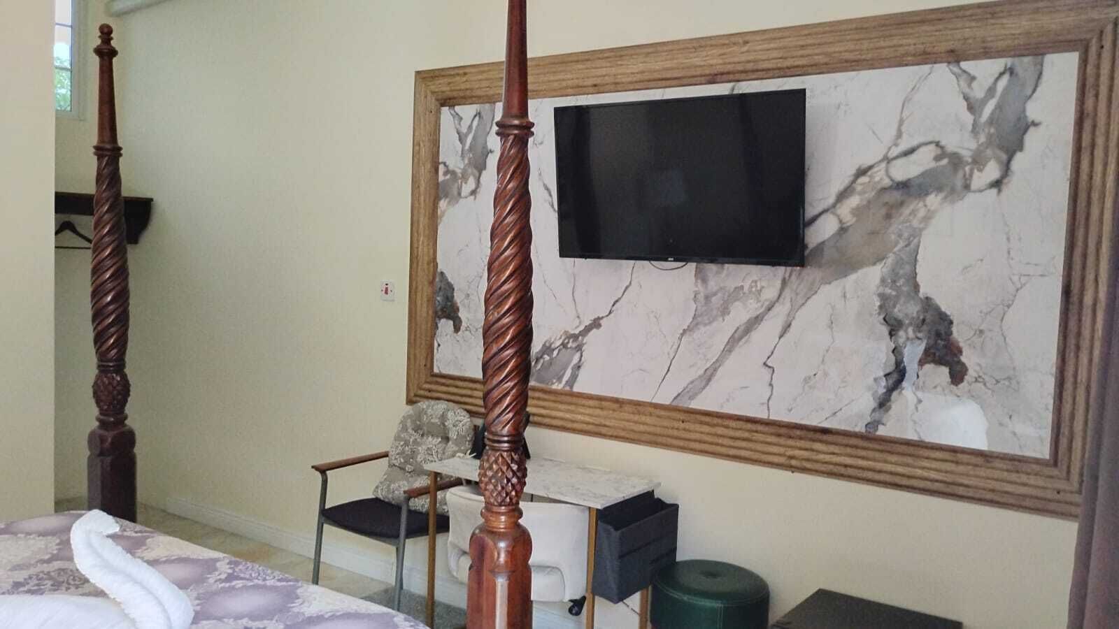 Un dormitorio con una televisión grande montada en una pared de mármol, una cama con cabecera de madera, una silla y una mesita.