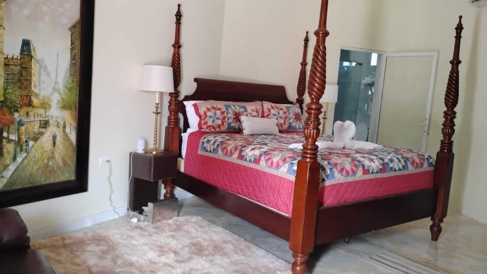 Een hemelbed met een bloemenquilt, een nachtkastje met een lamp en een schilderij aan de muur.