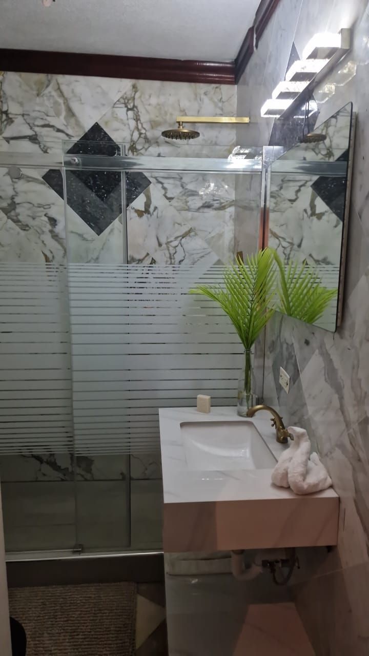 A bathroom with a glass shower enclosure, a marble backsplash, and a white sink with a towel and a plant.