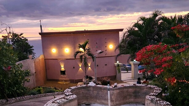 Un jardín ajardinado con un elemento circular de piedra, palmeras y un edificio con luces iluminadas al fondo.