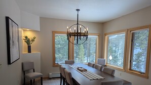A dining room with a long table, six chairs, a chandelier, large windows with a forest view, and a framed picture on the wall. - Quiet and Charming Big Sky home (Big Sky)