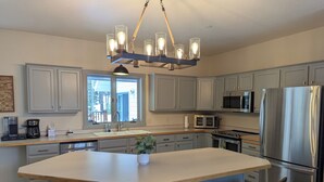 A modern kitchen with grey cabinets, a stainless steel refrigerator, a microwave, a coffee maker, and a central island with bar stools. - Quiet and Charming Big Sky home (Big Sky)