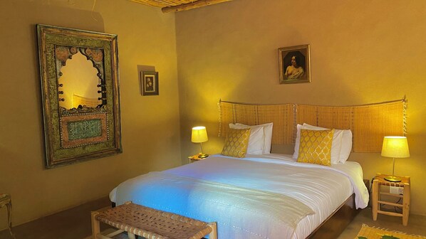 Habitación de hotel con una cama grande, dos lámparas de noche, un taburete de madera y una obra de arte enmarcada en la pared.