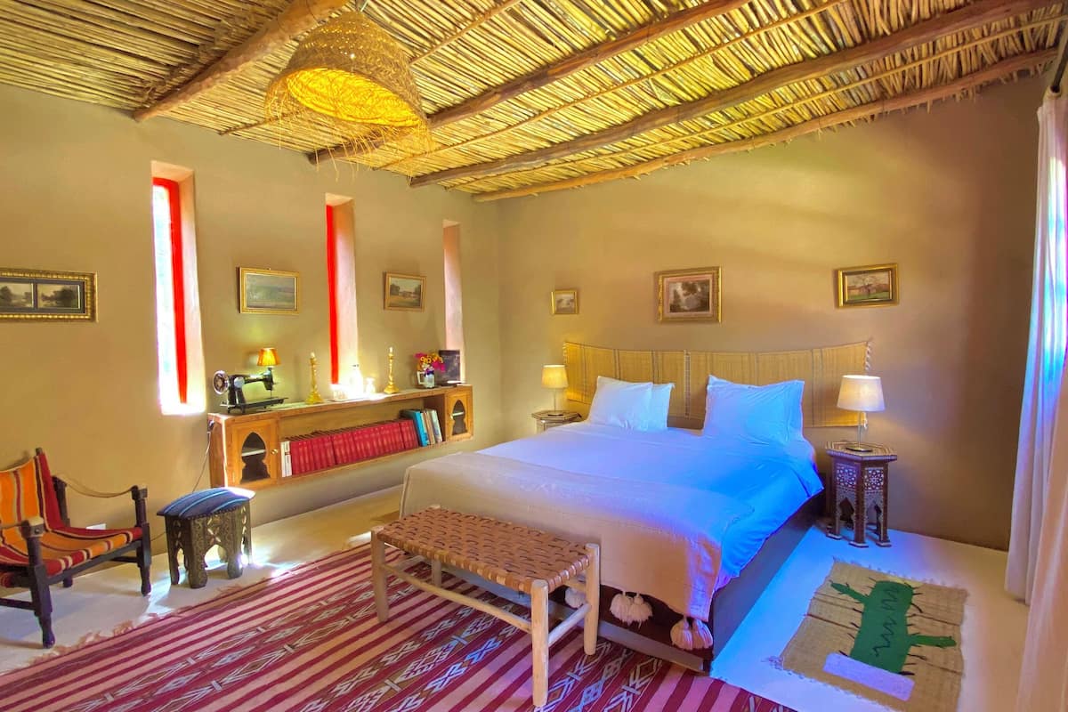 Una habitación con cama, una silla, un escritorio con máquina de coser y una ventana con cortinas rojas.