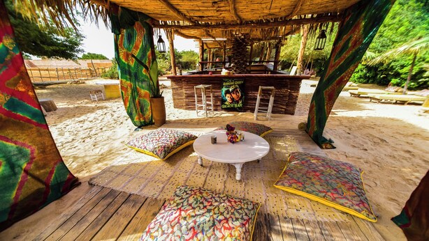 A beach bar area with a wooden deck, colorful cushions, a white table, and a bar counter. - Dar Tawarta (Dakhla)