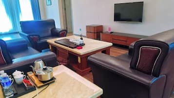 A hotel room with a sofa, a small table with a kettle, a flat-screen TV, and a window with curtains.
