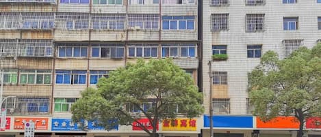 Un edificio de varios pisos con locales comerciales en la planta baja, con balcones y un árbol en primer plano.