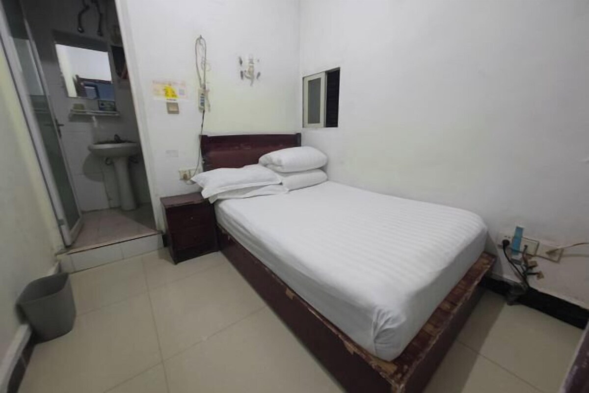 A small, clean room with a single bed, a bedside table, a small window, and a wall-mounted mirror.