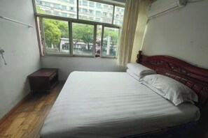 A single bed with a wooden headboard, a small bedside table, and a window with a view of buildings and trees. - Jingpai Hotel (Nanchang)