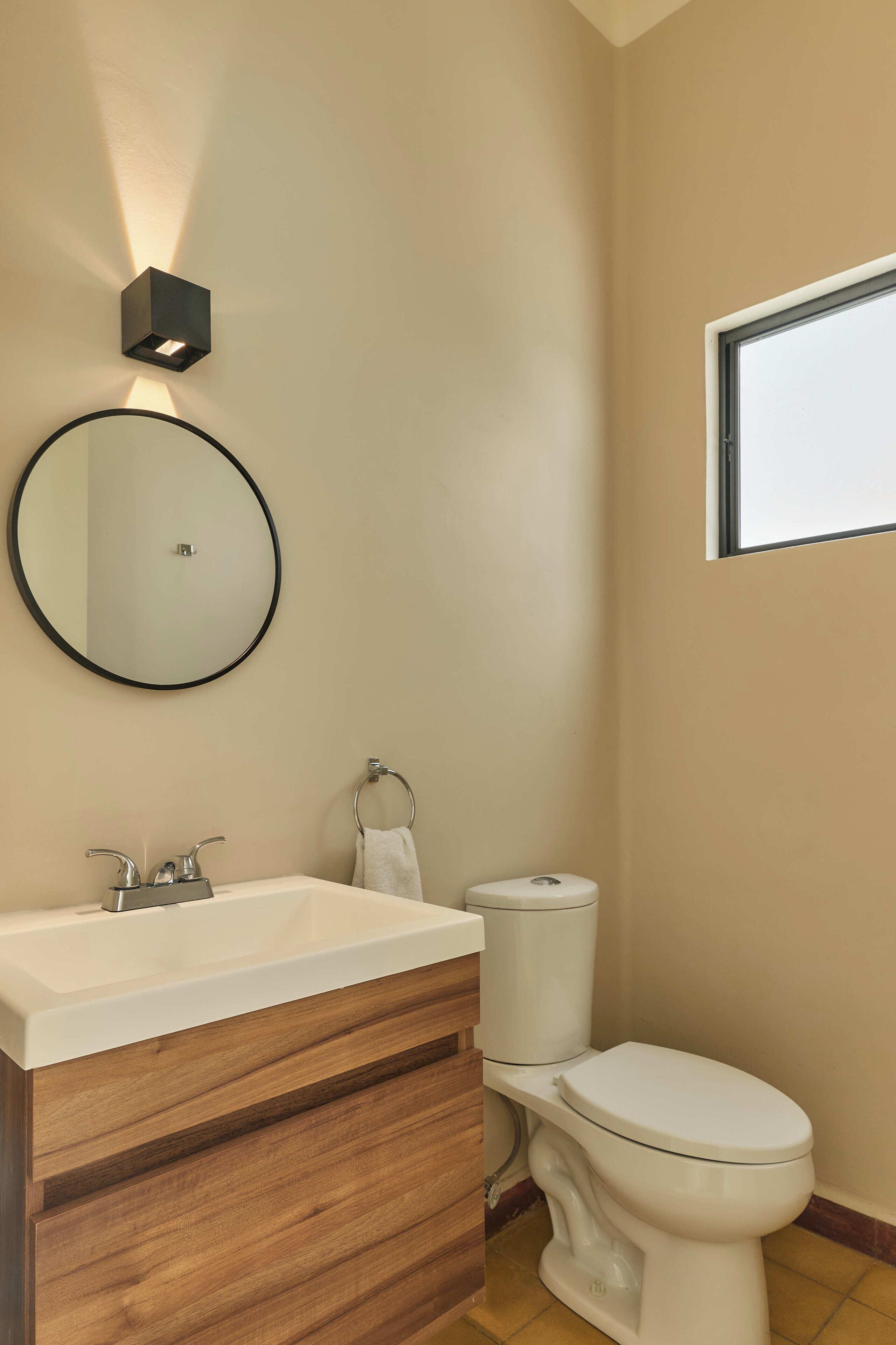 Un baño con tocador de madera, un espejo redondo, un inodoro y una lámpara de pared.