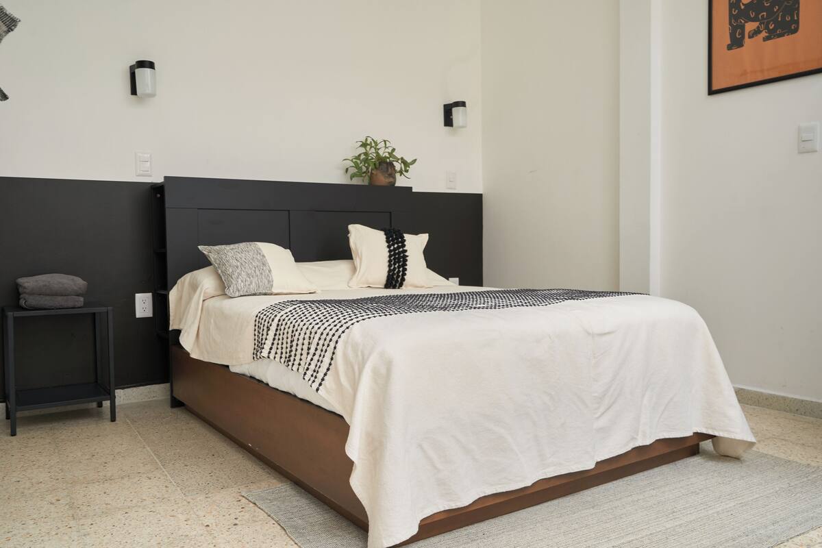 A neatly made bed with a white and black patterned bedspread, a dark headboard, and a small bedside table with a gray pillow.